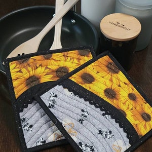 May include: Two quilted pot holders. One features a sunflower pattern with black trim and the other features a Mickey Mouse and Minnie Mouse pattern with black trim.