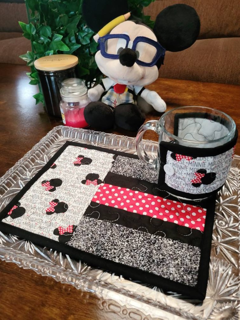 May include: A black and white quilted coaster with red polka dots and a Minnie Mouse design. The coaster is on a glass tray with a mug on top of it. The mug has a black and white Minnie Mouse design on it.