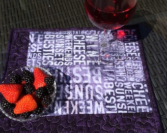 Könnte beinhalten: Lilafarbener gesteppter Platzdeckchen mit einem weißen Druck der Wörter "CHEESE", "WEEKENDS", "SUNSETS", "BESTIES" und mehr. Ein Glas Rotwein und eine Schüssel mit Erdbeeren und Brombeeren stehen auf dem Platzdeckchen.
