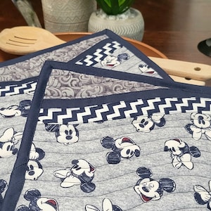 May include: Gray and navy blue quilted placemats featuring a repeating pattern of Mickey Mouse and Minnie Mouse. The placemats have a navy blue border and a decorative zigzag pattern.