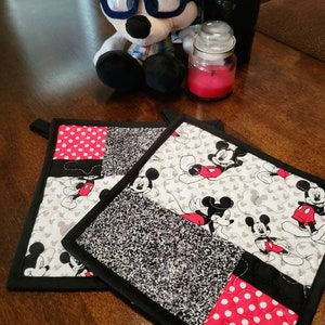 May include: Two black and white quilted pot holders with a Mickey Mouse theme. One pot holder has a black and white polka dot pattern, the other has a black and white speckled pattern. Both pot holders have a red and white Mickey Mouse design.