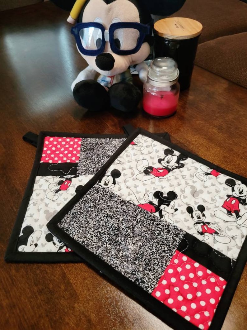 May include: Two black and white quilted pot holders with a Mickey Mouse theme. One pot holder has a black and white polka dot pattern, the other has a black and white speckled pattern. Both pot holders have a red and white Mickey Mouse design.