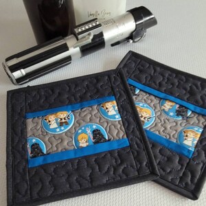 K&ouml;nnte beinhalten: Zwei schwarze, gesteppte Untersetzer aus Stoff mit einem blauen und grauen Stoffpanel mit einem Star Wars-Motiv mit Bildern von Darth Vader, Luke Skywalker und Prinzessin Leia. Die Untersetzer befinden sich auf einer wei&szlig;en Oberfl&auml;che.