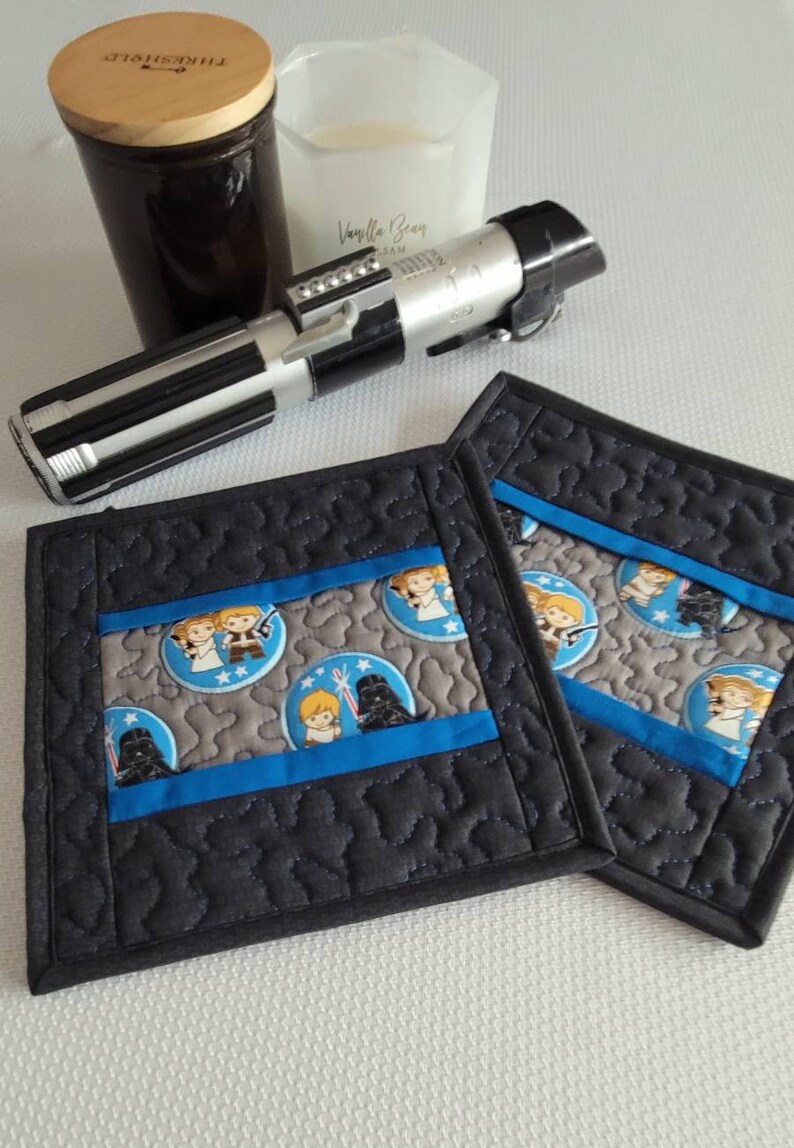 K&ouml;nnte beinhalten: Zwei schwarze, gesteppte Untersetzer aus Stoff mit einem blauen und grauen Stoffpanel mit einem Star Wars-Motiv mit Bildern von Darth Vader, Luke Skywalker und Prinzessin Leia. Die Untersetzer befinden sich auf einer wei&szlig;en Oberfl&auml;che.