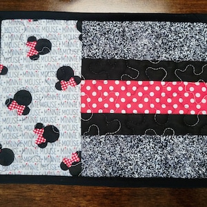 May include: A black and white quilted placemat with a red and white polka dot stripe. The placemat is decorated with Minnie Mouse silhouettes and the text "Minnie Mouse" repeated throughout.