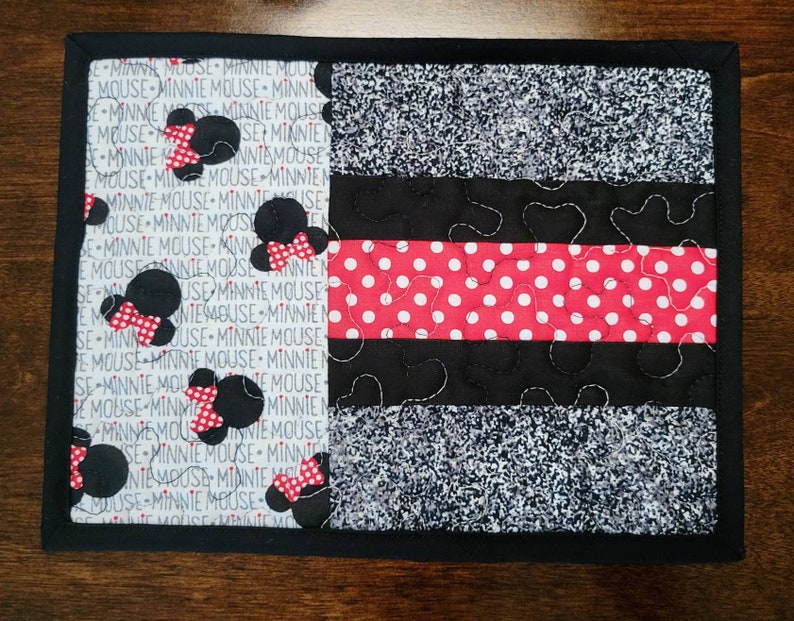 May include: A black and white quilted placemat with a red and white polka dot stripe. The placemat is decorated with Minnie Mouse silhouettes and the text "Minnie Mouse" repeated throughout.