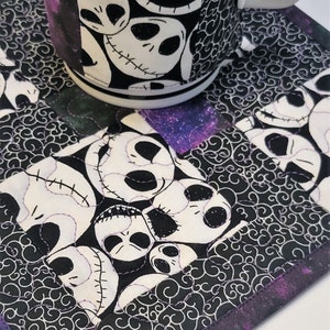 Puede incluir: Un posavasos de tela blanco y negro con un borde morado y negro presenta un patr&oacute;n repetido de la cara de Jack Skellington. Una taza blanca con una funda de tela blanco y negro con la cara de Jack Skellington est&aacute; sentada en el posavasos.