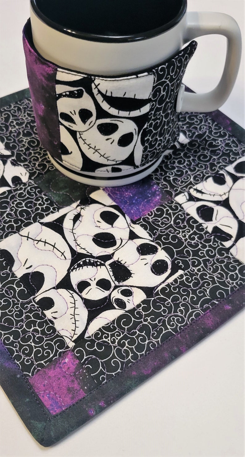 Puede incluir: Un posavasos de tela blanco y negro con un borde morado y negro presenta un patr&oacute;n repetido de la cara de Jack Skellington. Una taza blanca con una funda de tela blanco y negro con la cara de Jack Skellington est&aacute; sentada en el posavasos.