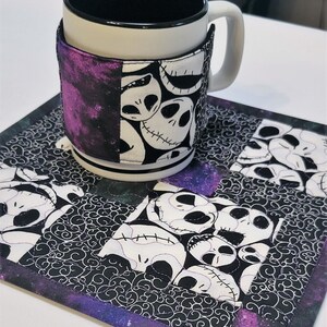 Puede incluir: Un posavasos acolchado en blanco y negro con un borde morado presenta un patr&oacute;n repetido de la cara de Jack Skellington de la pel&iacute;cula Pesadilla antes de Navidad. Una taza blanca con un borde negro y una funda de tela en blanco y negro con un fondo morado est&aacute; sentada en el posavasos.