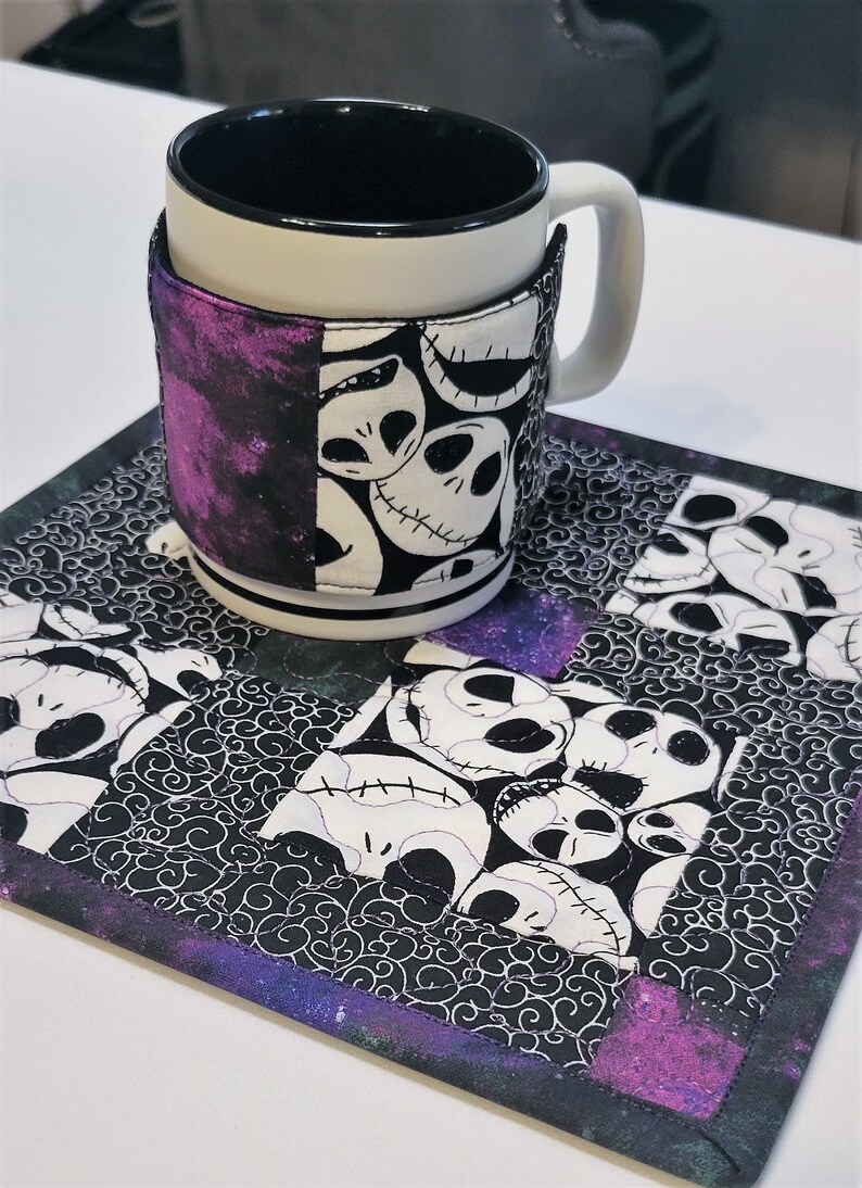Puede incluir: Un posavasos acolchado en blanco y negro con un borde morado presenta un patr&oacute;n repetido de la cara de Jack Skellington de la pel&iacute;cula Pesadilla antes de Navidad. Una taza blanca con un borde negro y una funda de tela en blanco y negro con un fondo morado est&aacute; sentada en el posavasos.