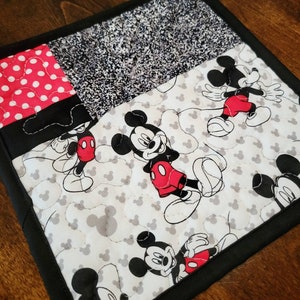 May include: A black and white quilted hot pad with a Mickey Mouse design. The hot pad features a patchwork design with a red and white polka dot fabric, a black and white speckled fabric, and a white fabric with Mickey Mouse heads printed on it. The hot pad has a black border.