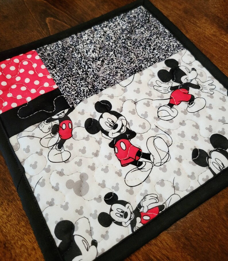 May include: A black and white quilted hot pad with a Mickey Mouse design. The hot pad features a patchwork design with a red and white polka dot fabric, a black and white speckled fabric, and a white fabric with Mickey Mouse heads printed on it. The hot pad has a black border.