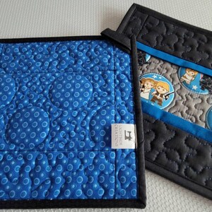 K&ouml;nnte beinhalten: Zwei gequiltete Topflappen mit Star Wars-Motiv. Ein Topflappen ist blau mit wei&szlig;en Punkten und der andere ist schwarz mit grauen Sternen. Beide Topflappen haben ein blaues und graues Stoffpanel mit Bildern von Star Wars-Charakteren.