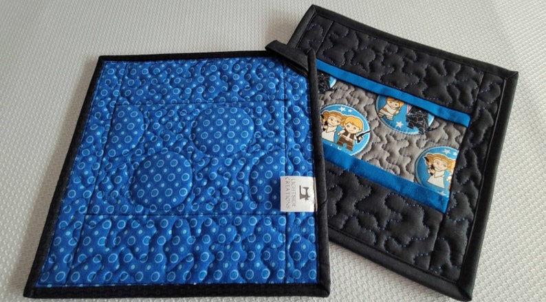 K&ouml;nnte beinhalten: Zwei gequiltete Topflappen mit Star Wars-Motiv. Ein Topflappen ist blau mit wei&szlig;en Punkten und der andere ist schwarz mit grauen Sternen. Beide Topflappen haben ein blaues und graues Stoffpanel mit Bildern von Star Wars-Charakteren.