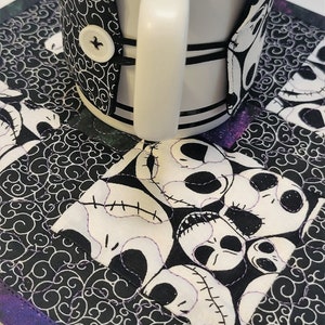Puede incluir: Una taza de cer&aacute;mica blanca con un calientavasos de tela blanco y negro con un patr&oacute;n repetido de la cara de Jack Skellington de Pesadilla antes de Navidad. El calientavasos est&aacute; sobre un posavasos de tela blanco y negro con un borde morado y negro.