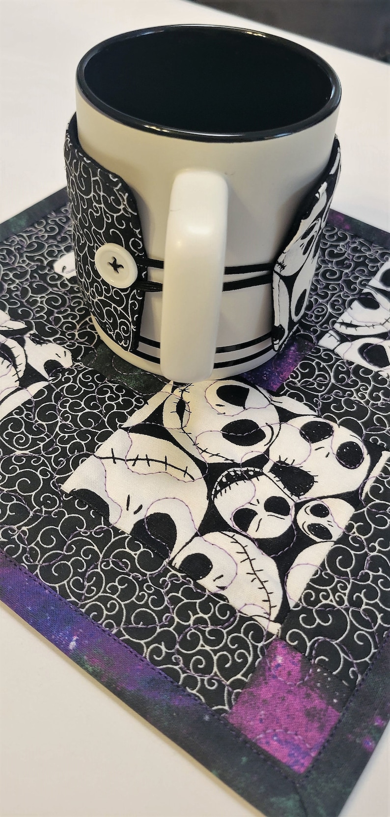 Puede incluir: Una taza de cer&aacute;mica blanca con un calientavasos de tela blanco y negro con un patr&oacute;n repetido de la cara de Jack Skellington de Pesadilla antes de Navidad. El calientavasos est&aacute; sobre un posavasos de tela blanco y negro con un borde morado y negro.
