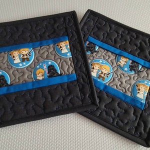 May include: Two black quilted pot holders with a blue and gray Star Wars themed fabric insert. The fabric features characters from Star Wars, including Darth Vader, Luke Skywalker, and Princess Leia.