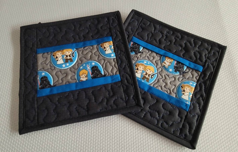 K&ouml;nnte beinhalten: Zwei schwarze, gesteppte Topflappen mit einem blauen und grauen Stoff-Einsatz im Star Wars-Design. Der Stoff zeigt Figuren aus Star Wars, darunter Darth Vader, Luke Skywalker und Prinzessin Leia.