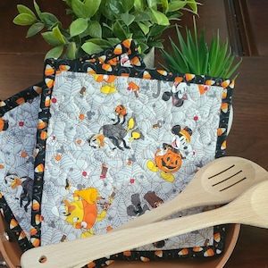 May include: Two black and orange Halloween themed pot holders with a white background featuring Mickey Mouse and other characters. The pot holders are on top of a brown pan with two wooden spoons.