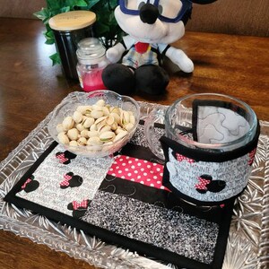 May include: A black and white quilted coaster with red polka dots and the repeated text "Minnie Mouse" surrounding a black silhouette of Minnie Mouse's head with a red bow. A glass mug with a black and white patterned sleeve sits on the coaster. A small bowl filled with pistachios sits on a clear glass tray.