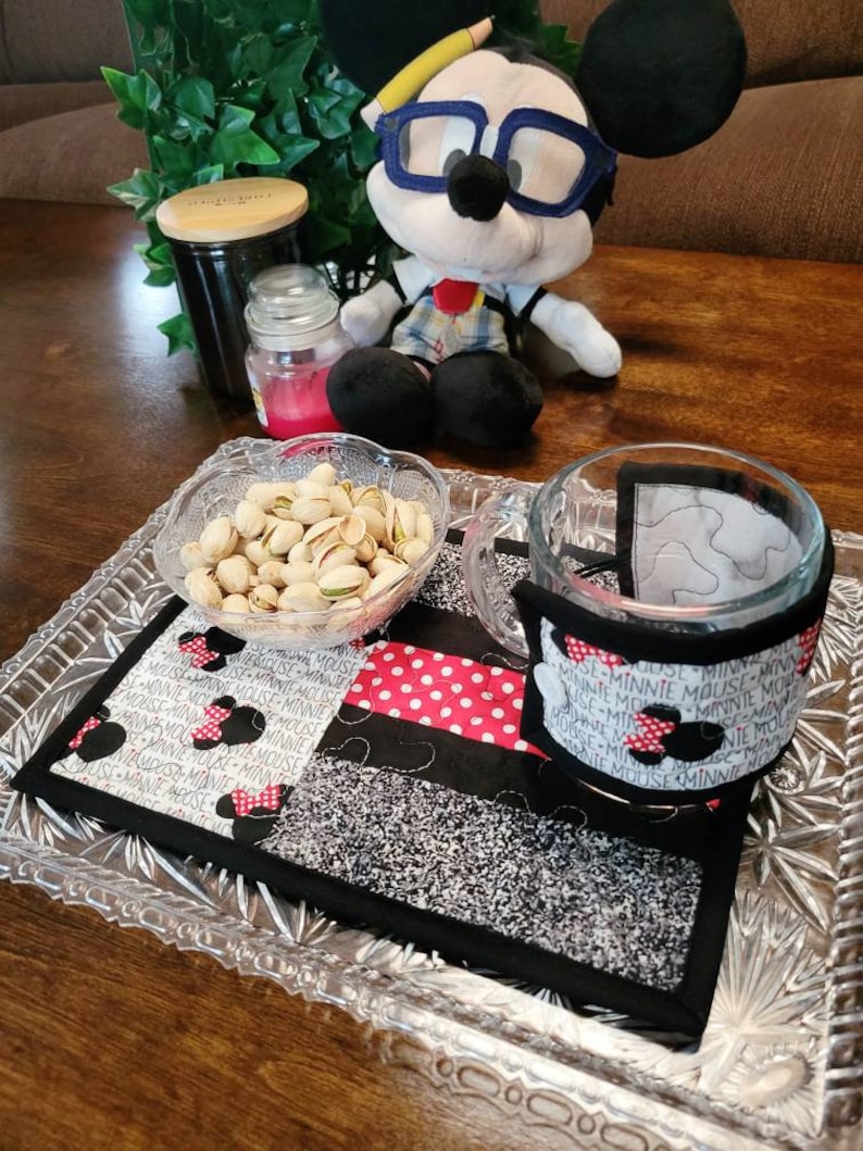 May include: A black and white quilted coaster with red polka dots and the repeated text "Minnie Mouse" surrounding a black silhouette of Minnie Mouse's head with a red bow. A glass mug with a black and white patterned sleeve sits on the coaster. A small bowl filled with pistachios sits on a clear glass tray.