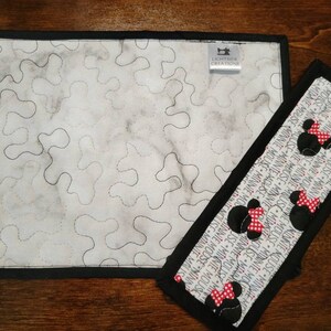 May include: A white quilted placemat with a black border and a black and white Minnie Mouse print fabric hot pad. The hot pad has a black border and a white background with repeated images of Minnie Mouse with red bows. The placemat has a label that reads "Lightslide Creations".
