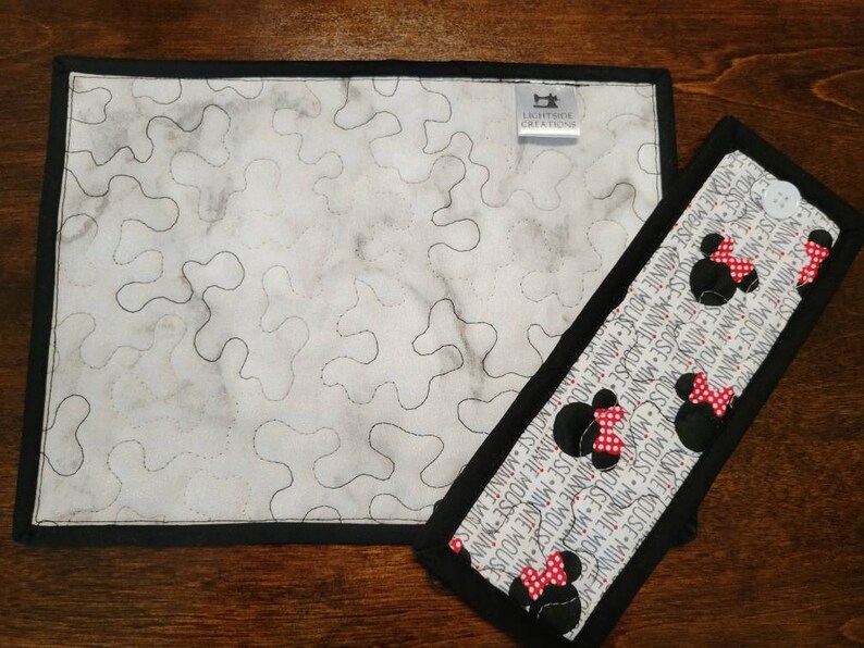May include: A white quilted placemat with a black border and a black and white Minnie Mouse print fabric hot pad. The hot pad has a black border and a white background with repeated images of Minnie Mouse with red bows. The placemat has a label that reads "Lightslide Creations".
