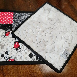 May include: Two black and white quilted pot holders with a Mickey Mouse design. One pot holder has a red and white polka dot square. The other pot holder has a white background with a black quilted pattern. The pot holders have black fabric trim and a black hanging loop.