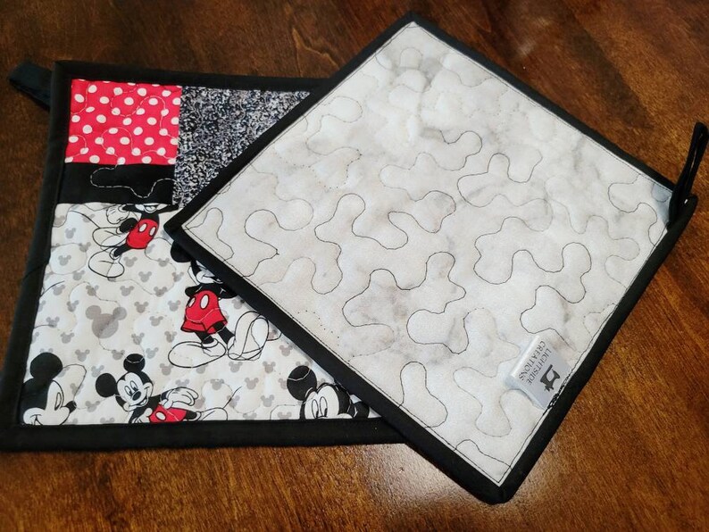 May include: Two black and white quilted pot holders with a Mickey Mouse design. One pot holder has a red and white polka dot square. The other pot holder has a white background with a black quilted pattern. The pot holders have black fabric trim and a black hanging loop.