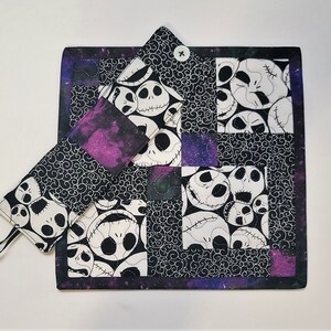Puede incluir: Un posavasos de tela acolchado en blanco y negro con un patr&oacute;n repetido de la cara de Jack Skellington de la pel&iacute;cula Pesadilla antes de Navidad. El posavasos tiene un borde morado y negro y un bot&oacute;n en el centro.