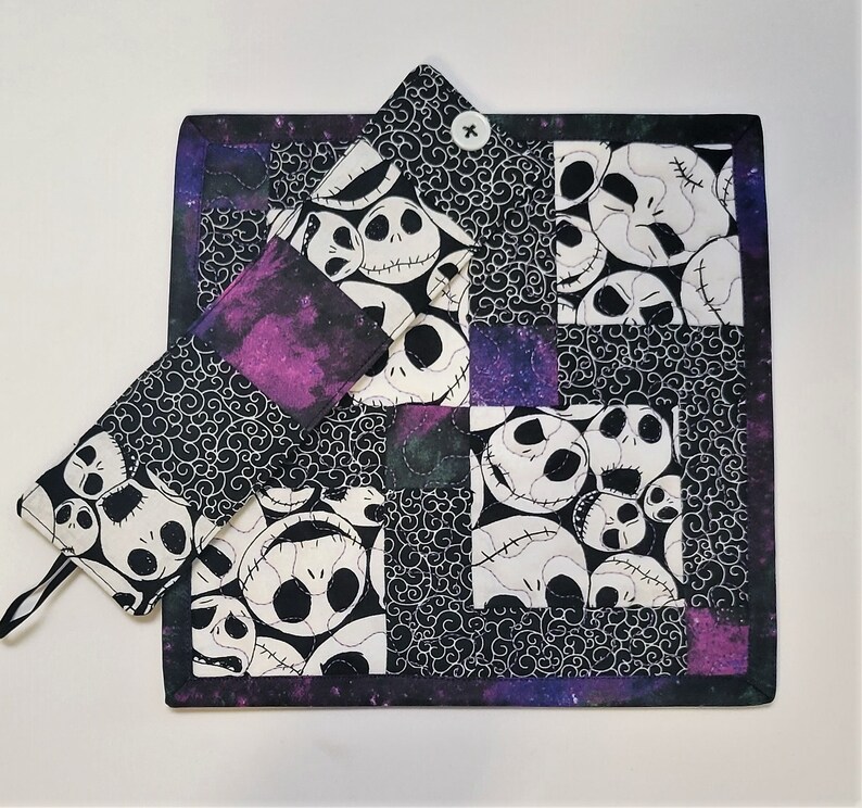 Puede incluir: Un posavasos de tela acolchado en blanco y negro con un patr&oacute;n repetido de la cara de Jack Skellington de la pel&iacute;cula Pesadilla antes de Navidad. El posavasos tiene un borde morado y negro y un bot&oacute;n en el centro.