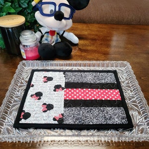 May include: A black and white fabric placemat with a red and white polka dot stripe and the text "Minnie Mouse" repeated throughout. The placemat is on a glass tray with a crystal pattern.
