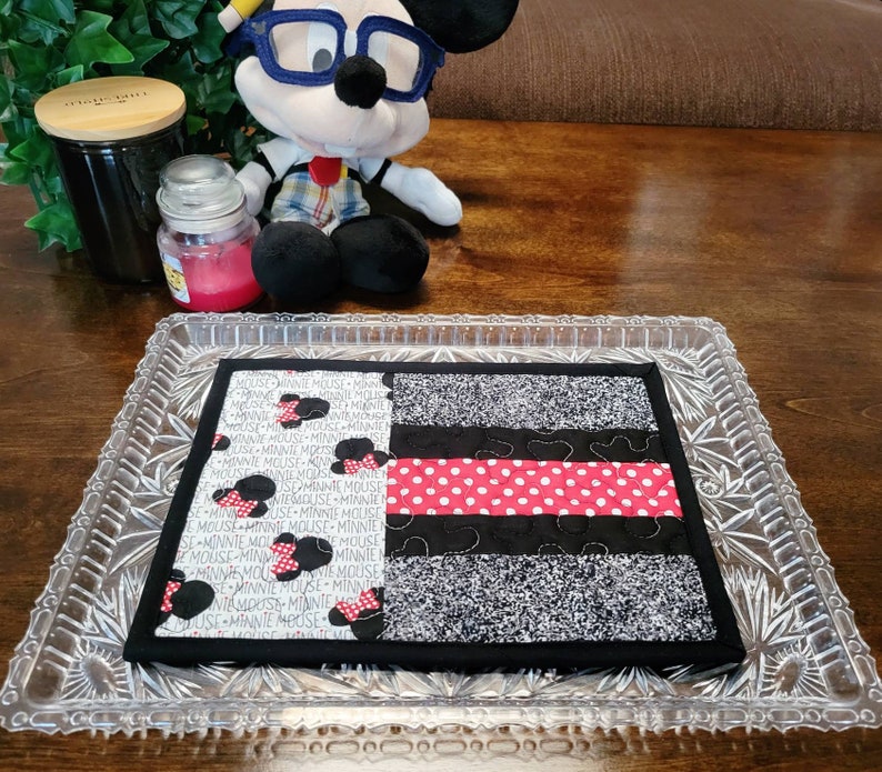 May include: A black and white fabric placemat with a red and white polka dot stripe and the text "Minnie Mouse" repeated throughout. The placemat is on a glass tray with a crystal pattern.