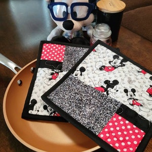 May include: Two black and red quilted pot holders with a Mickey Mouse pattern. The pot holders are on top of a brown frying pan.
