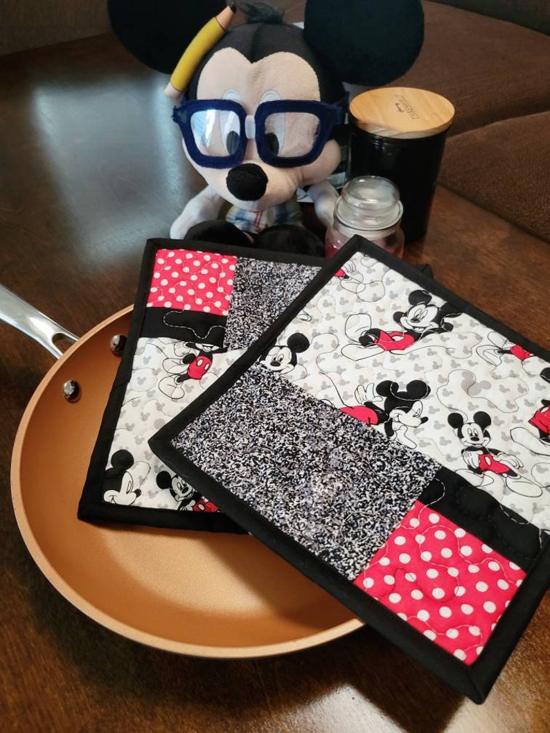 May include: Two black and red quilted pot holders with a Mickey Mouse pattern. The pot holders are on top of a brown frying pan.