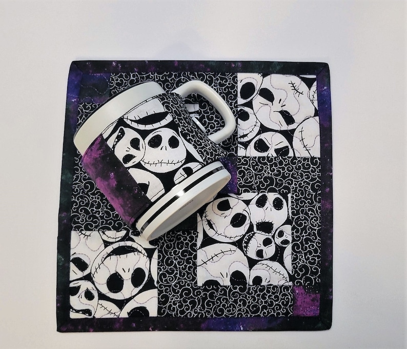 Puede incluir: Una taza blanca y negra con un patr&oacute;n del rostro de Jack Skellington de la pel&iacute;cula Pesadilla antes de Navidad. La taza est&aacute; sobre un posavasos de tela blanco y negro con un patr&oacute;n similar. El posavasos tiene un borde morado y negro.