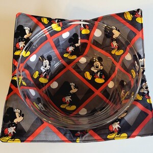 May include: A clear glass bowl rests on a square, padded bowl cosy. The cosy has a repeating pattern of Mickey Mouse in various poses, set against a grey background with red grid lines. The bowl is empty.
