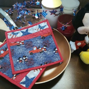 May include: Two red, white and blue quilted pot holders with a Mickey Mouse and fireworks design. The pot holders are on a copper-coloured plate. A Mickey Mouse plush toy is in the background.