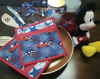 Könnte beinhalten: Zwei rote, weiße und blaue gequiltete Topflappen mit einem Mickey-Maus- und Feuerwerksmotiv. Die Topflappen liegen auf einem kupferfarbenen Teller. Ein Plüschtier Mickey Mouse befindet sich im Hintergrund.
