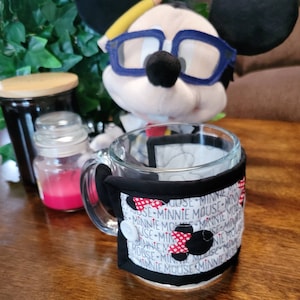 May include: A black and white fabric mug cozy with red polka dots and the text "Minnie Mouse" repeated around the cozy. The cozy is on a glass mug with a handle.