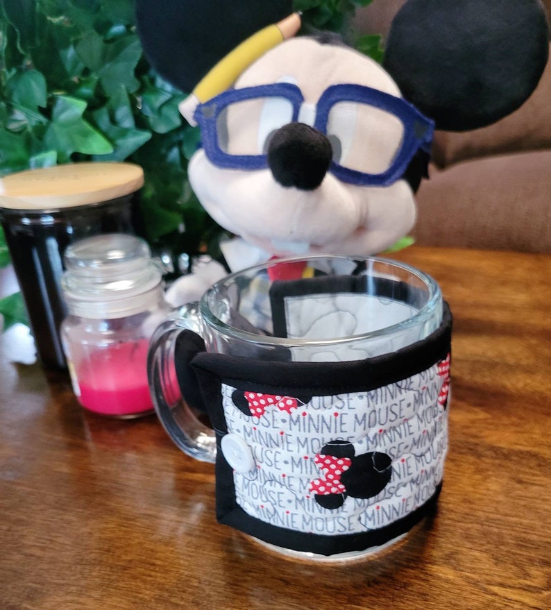 May include: A black and white fabric mug cozy with red polka dots and the text "Minnie Mouse" repeated around the cozy. The cozy is on a glass mug with a handle.