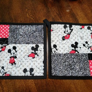 May include: Two black and red quilted pot holders with a Mickey Mouse design. The pot holders have a black border and are made of different fabrics, including red with white polka dots, black and white speckled fabric, and white fabric with a Mickey Mouse pattern.