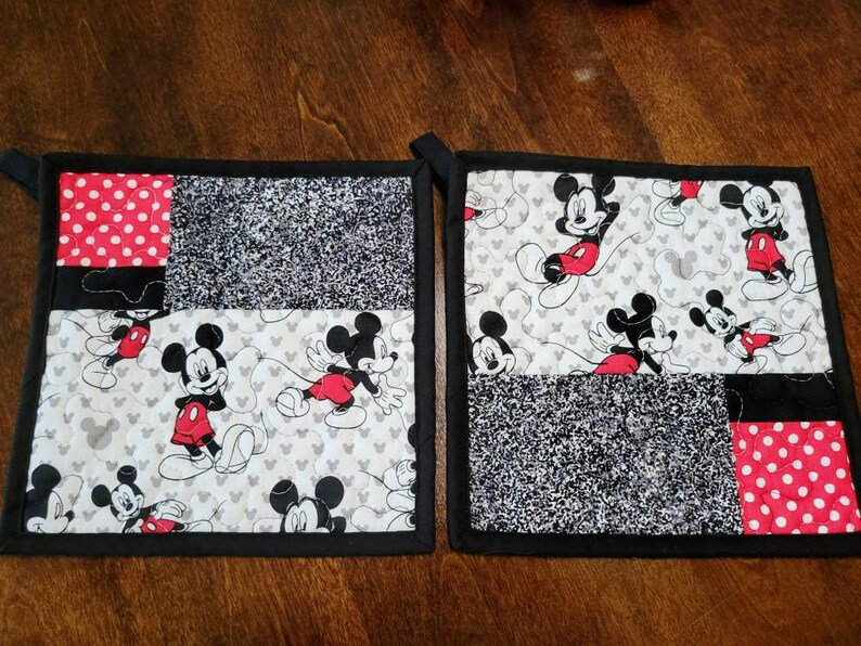 May include: Two black and red quilted pot holders with a Mickey Mouse design. The pot holders have a black border and are made of different fabrics, including red with white polka dots, black and white speckled fabric, and white fabric with a Mickey Mouse pattern.