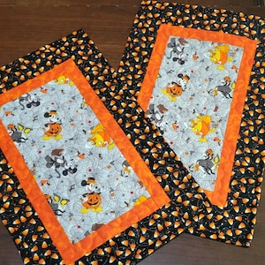Könnte beinhalten: Zwei Tischläufer zum Thema Halloween mit einem schwarzen und orangefarbenen Mais-Muster. Die Mitte der Läufer besteht aus weißem Stoff mit Mickey Mouse und anderen Figuren in einer Halloween-Szene.