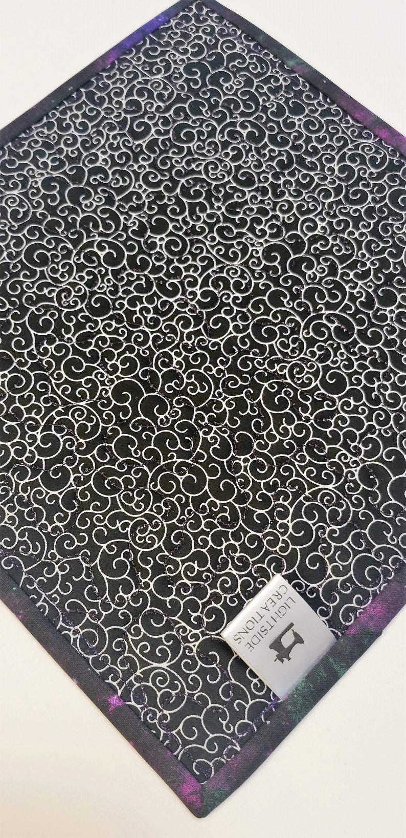 Puede incluir: Tela negra y blanca con un patr&oacute;n de espiral. La tela tiene un borde morado, verde y negro. Una etiqueta plateada con el texto "LIGHT SIDE CREATIONS" y un icono de m&aacute;quina de coser est&aacute; unida a la tela.