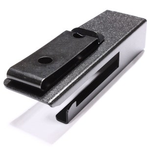 May include: A dark gray metal clip with a textured finish. The clip has a rectangular body with a slot and a hinged, spring-loaded clip. The clip is designed to hold items securely.
