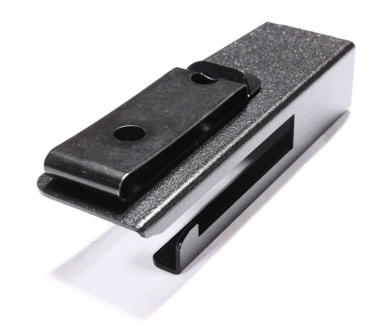 May include: A dark gray metal clip with a textured finish. The clip has a rectangular body with a slot and a hinged, spring-loaded clip. The clip is designed to hold items securely.
