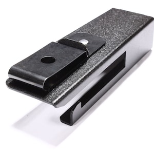 May include: A dark gray metal clip with a textured finish. The clip has a rectangular body with a hinged, curved top piece and a slot on the side. The design is simple and industrial.