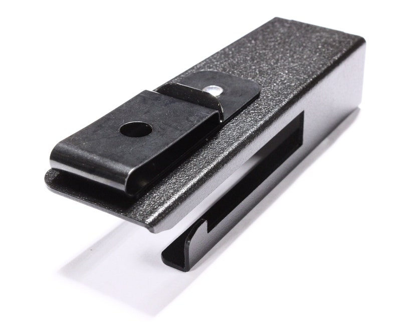 May include: A dark gray metal clip with a textured finish. The clip has a rectangular body with a hinged, curved top piece and a slot on the side. The design is simple and industrial.