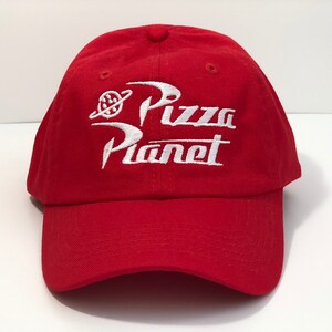 May include: A red baseball cap with white embroidered "Pizza Planet" text. The cap has a curved peak and a small button on top. The design includes a planet and a ring, a classic cap style.